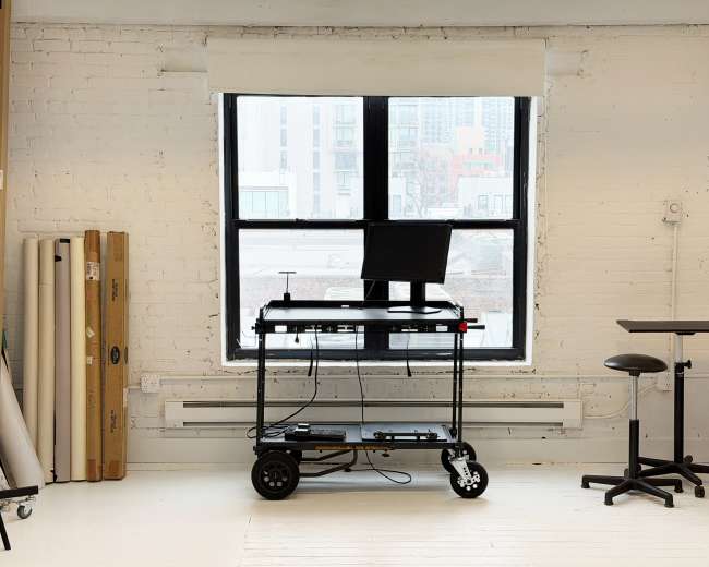 A black adjustable-height desk with a computer monitor is positioned in front of a large window in a spacious, minimalist room with white brick walls.