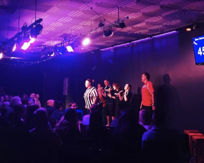 A group of performers is on stage in a dimly lit theater while an audience watches from seated rows.