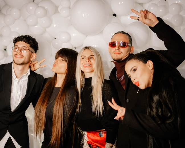 A group of five individuals poses with peace signs in front of a backdrop of large white balloons.