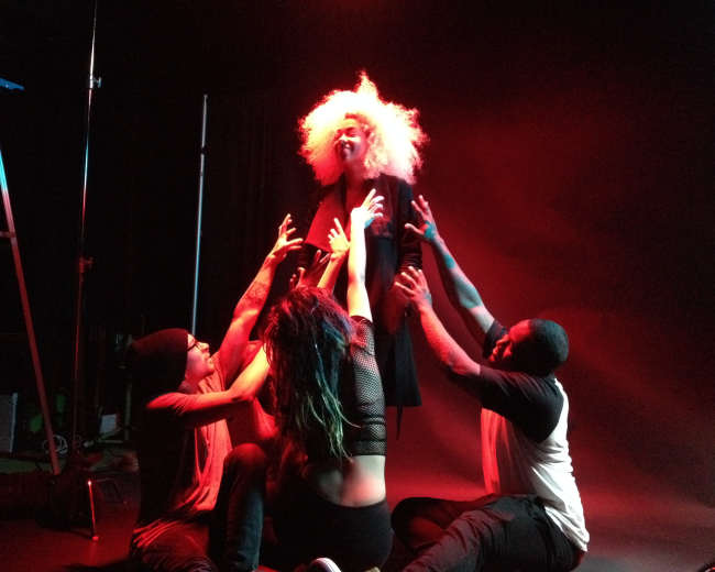 A person with an elaborate hairstyle is surrounded by four individuals, who are reaching towards them on a stage with dramatic lighting.