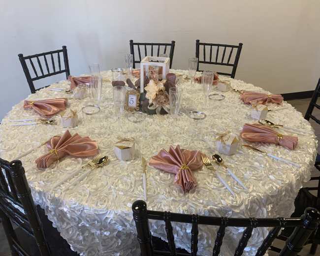 A round table is set with a white floral tablecloth, elegant place settings, and decorative napkins in a banquet room.