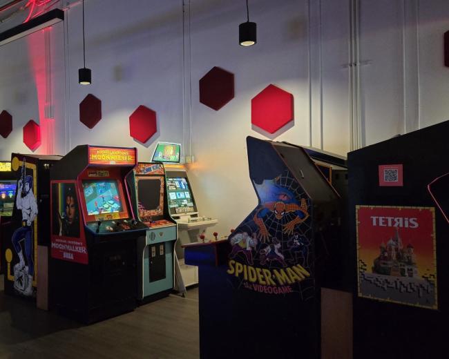 The image shows a row of vintage arcade machines displayed against a white wall with red hexagonal accents, illuminated in dim lighting.
