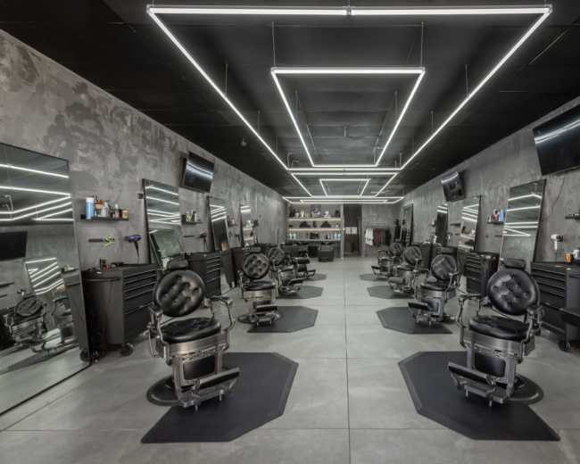 The image shows a modern barber shop with several black barber chairs arranged in a spacious, well-lit interior featuring mirrored walls and sleek lighting.