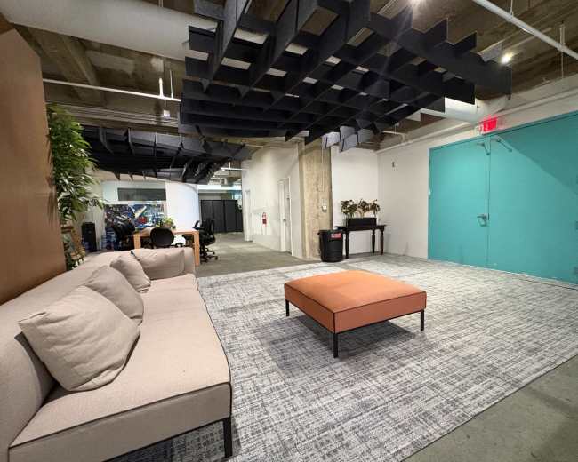The image shows a modern office lounge area with a beige sofa, an orange coffee table, and a textured gray carpet, complemented by a vibrant turquoise door.