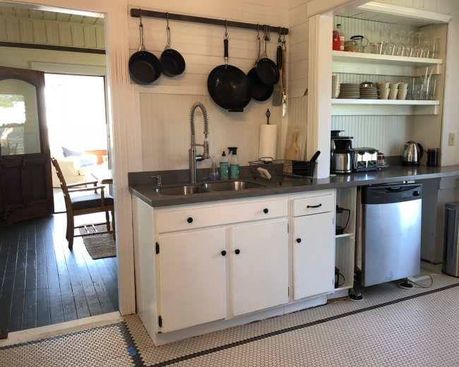 The image shows a small kitchen with a stainless steel countertop, white cabinets, hanging pots, and an array of kitchen appliances.