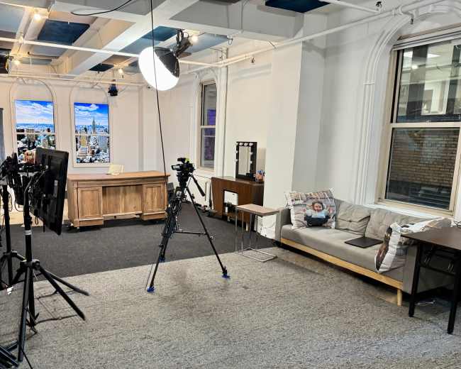 The image shows a bright studio space with a wooden desk, a sofa, and several cameras set up for filming.