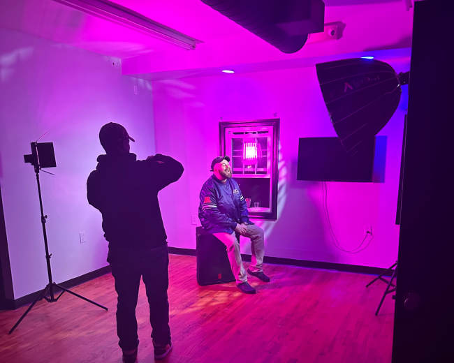 A person sits on a stool in a brightly lit studio with purple lighting, while another person photographs them.