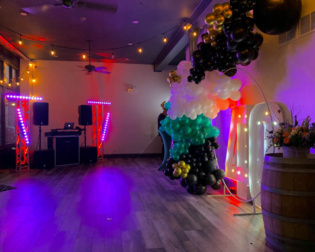 The interior of a venue is decorated with colorful balloons and lights, featuring a DJ setup on one side and a large floral arrangement on a barrel.