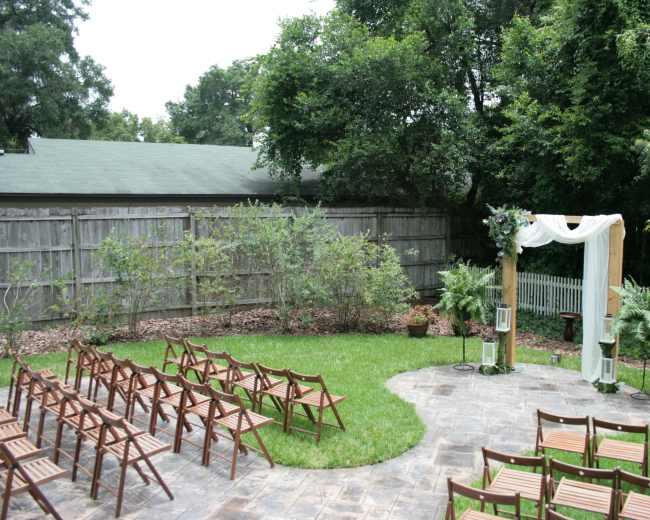 A wedding ceremony setup features wooden chairs arranged in rows facing a decorated archway surrounded by greenery in a garden area.