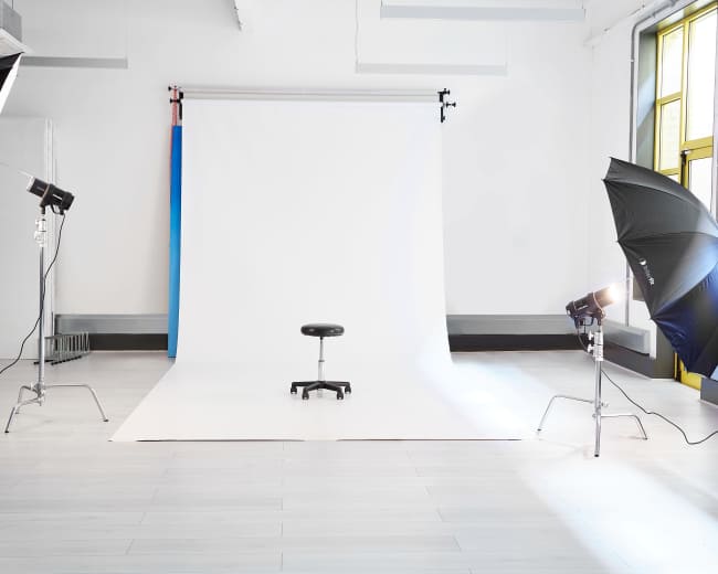 A photography studio features a white backdrop with a stool in the center, illuminated by two softbox lights.