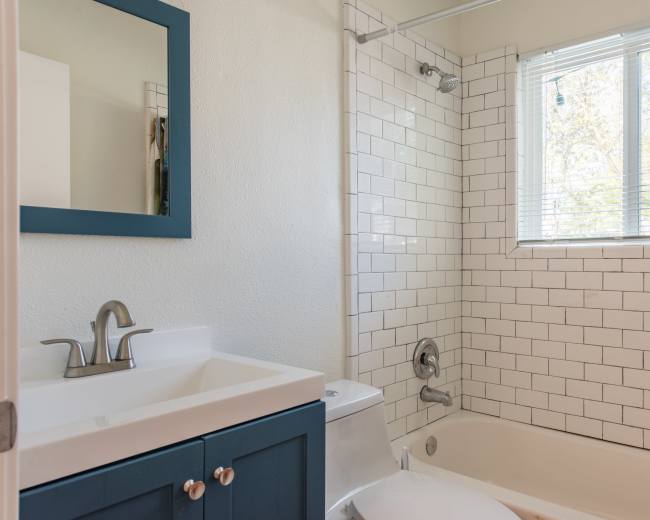 A bright bathroom featuring a white tiled shower area, a bathtub, a toilet, and a blue-framed mirror above a sink with a dark blue cabinet.