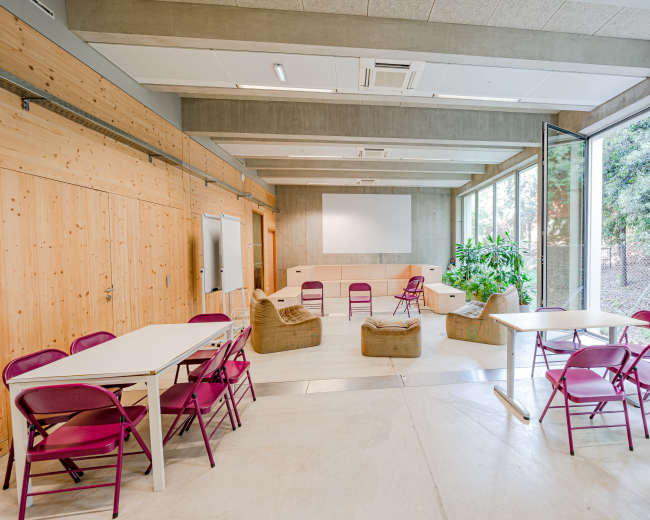 A modern classroom with several tables and chairs arranged around a central seating area, surrounded by large windows and greenery outside.