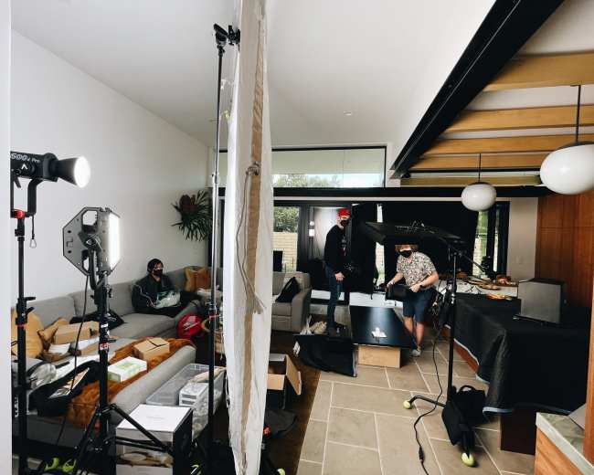 A cluttered living room set up for a photoshoot, featuring various equipment, two people wearing masks, and bright lighting.
