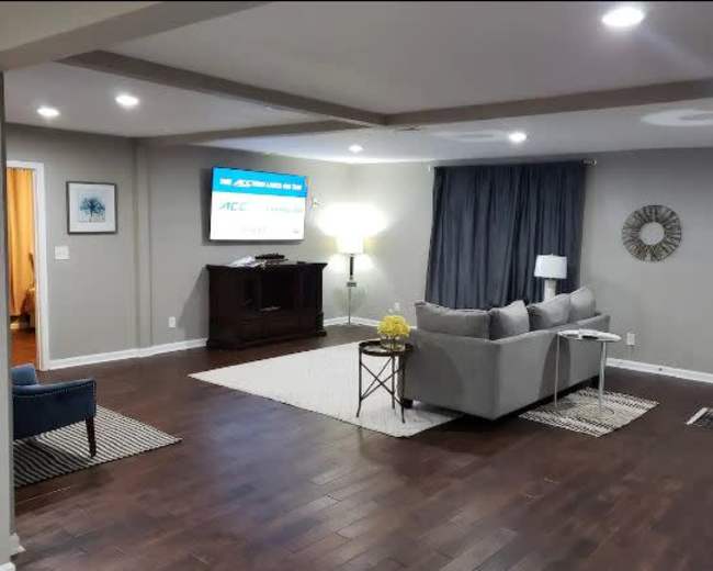 The living room features a gray sofa, a coffee table, a dark wooden cabinet, and a large screen mounted on the wall, with warm lighting and a mix of hardwood and rug flooring.