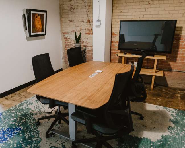 A modern conference room features a wooden table surrounded by five black chairs, a television mounted on the wall, and brick accents.