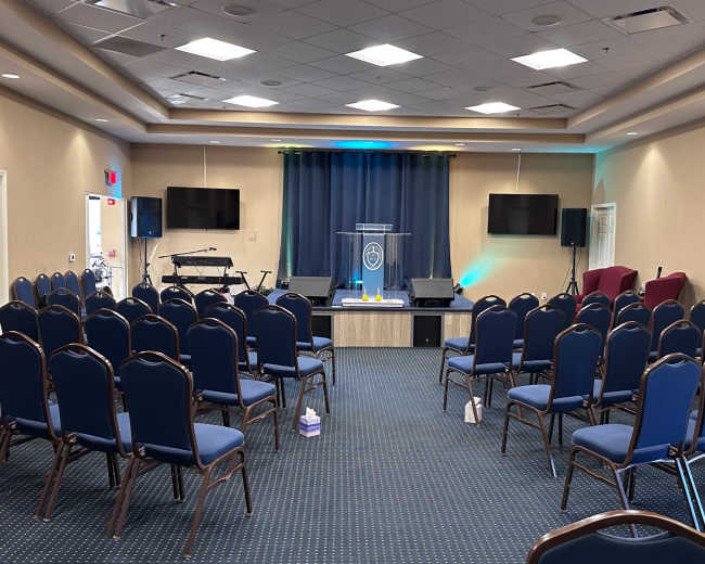 A spacious church interior with rows of blue chairs facing a stage that has a podium and two television screens.