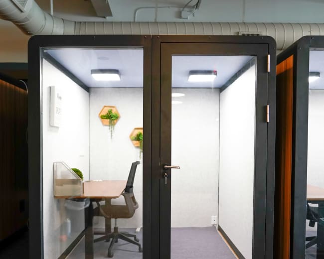 The image shows a glass-walled office pod with a desk and chair inside, situated in an office environment.