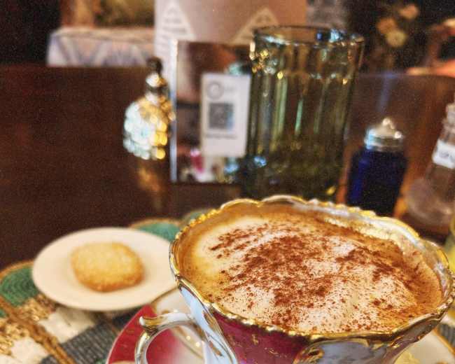 A beautifully designed cup of coffee topped with cocoa sits on a decorative coasters next to a small cookie.