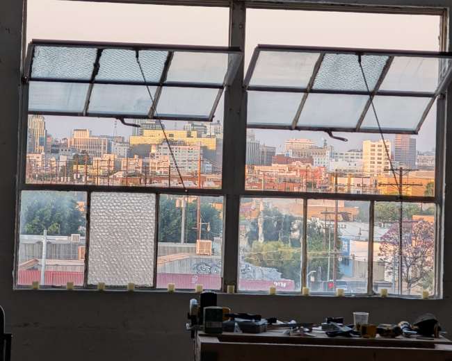 The image shows a workshop interior with a large window offering a view of a city skyline at sunset.