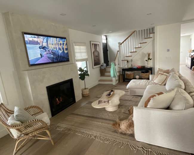 A modern living room features a white sectional sofa, a coffee table, a fireplace, and a television mounted on the wall, with stairs leading to an upper level.