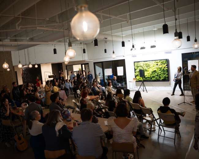 A crowd gathers in a modern event space with hanging lights, as a speaker addresses the audience near a large green wall display.