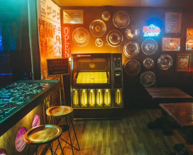 The image shows a dimly lit bar interior featuring a vintage jukebox, wooden tables, and a decorative wall adorned with various framed memorabilia.