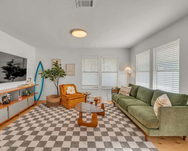 The living room features a green sofa, a mustard yellow armchair, a coffee table, and a large television against a white wall, with a checkered rug and house plants adding to the decor.
