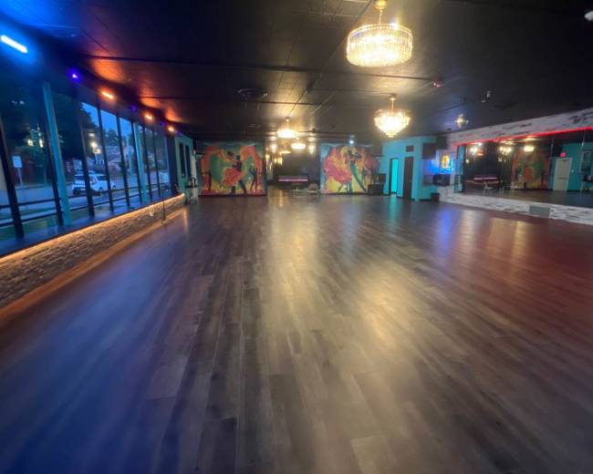 The image shows a dance studio with wooden flooring, large windows, colorful wall murals, and overhead chandeliers.