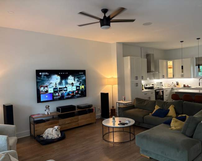 The image shows a modern living room with a large TV mounted on the wall, a green sectional sofa, a coffee table, and a view of a kitchen in the background.