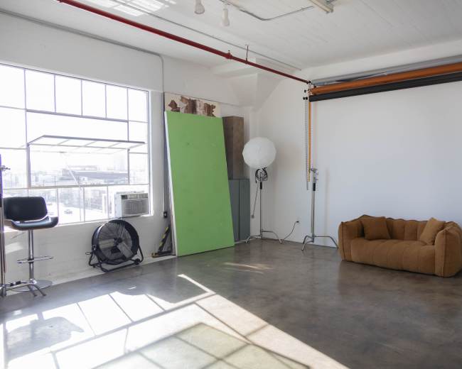 The image shows an empty studio space with large windows, a couch, and various photography equipment set against white walls.