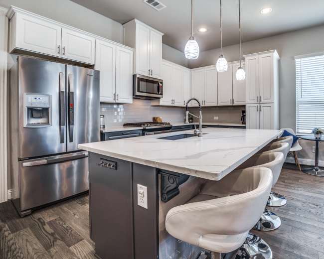 The image shows a modern kitchen featuring white cabinetry, stainless steel appliances, a marble-topped island with bar stools, and a cozy seating area with blue accents.