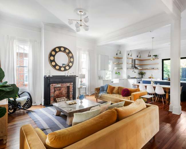 The open living space features a comfortable seating area with yellow sofas, a fireplace with a large round mirror above it, and a modern kitchen visible in the background.