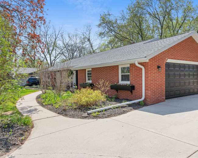 A brick house with a black garage door is surrounded by landscaped greenery and a curved concrete driveway.