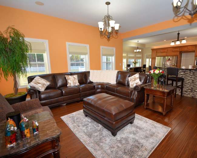 The living room features a brown leather sectional sofa, a coffee table, and a kitchen in the background with wooden cabinetry.