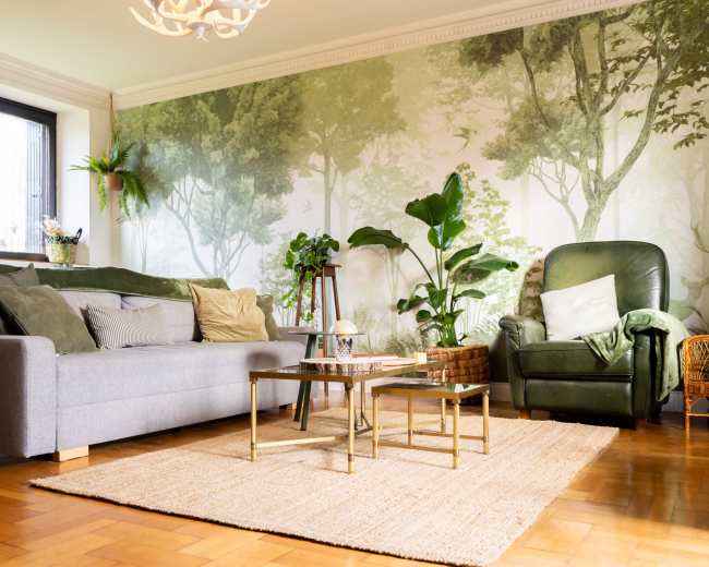 A cozy living room features a gray sofa and a green armchair, surrounded by plants and a large wall mural of trees.