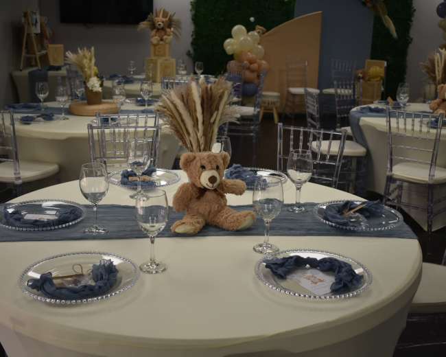 A decorated banquet hall features round tables with white tablecloths, each set with crystal glassware and a plush teddy bear centerpiece.
