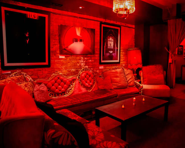 The image shows a dimly lit lounge area featuring red walls, several framed artworks, and plush seating arrangements.