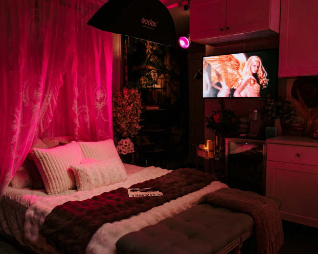 A bedroom is decorated with pink lighting, featuring a large bed with plush bedding and a television displaying an image of a woman.