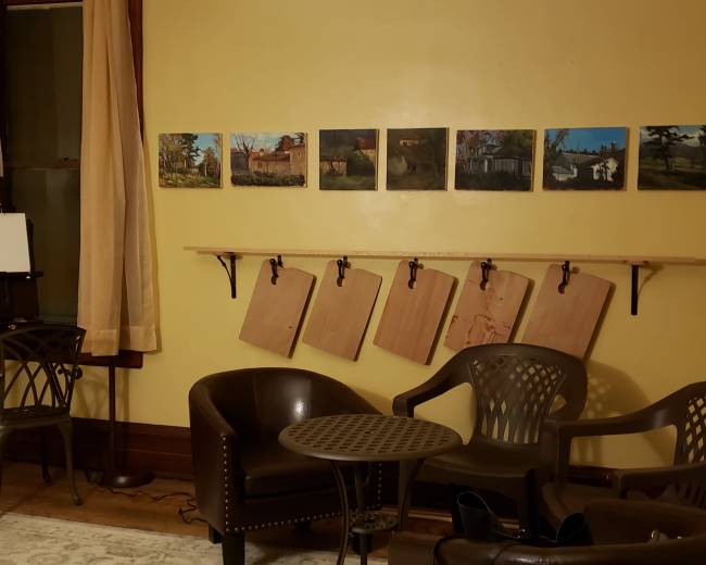An artist's studio with chairs, a small table, and paintings displayed on the wall above hung wooden boards.