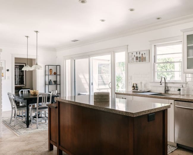A modern kitchen features a central island with a granite countertop, stainless steel appliances, and white cabinetry, leading to a dining area with a wooden table and metal chairs.
