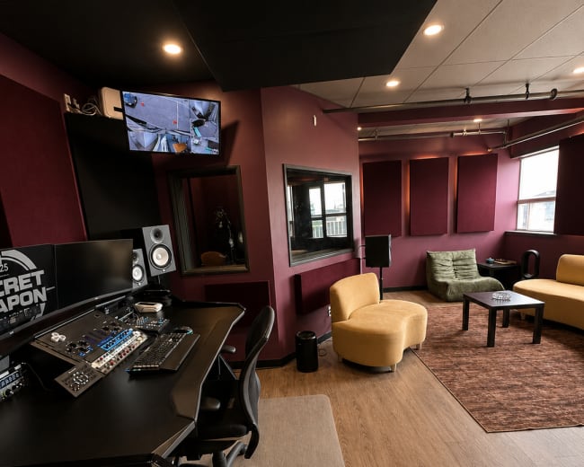 The image shows a modern recording studio featuring a control desk, comfortable seating, and soundproofed walls in a well-lit space.