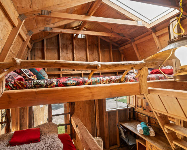 The image shows the interior of a rustic wooden cabin featuring a lofted sleeping area with colorful bedding and a cozy ground level with a plush bed and a small table.