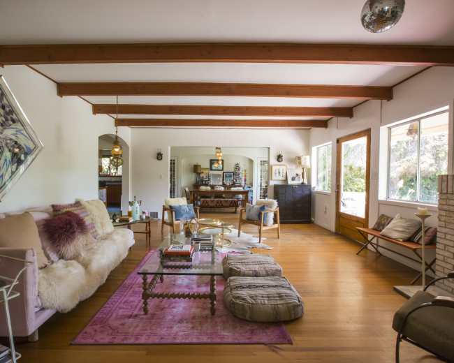 The living room features wooden beams, a plush sofa, a coffee table with decorative items, and large windows providing natural light.