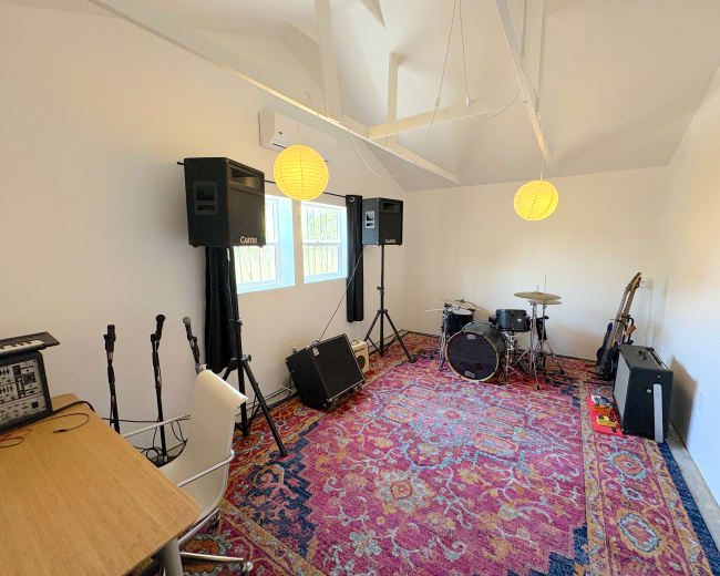 The room features a music setup with a drum kit, amplifiers, microphones, and a desk, all arranged on a patterned rug.