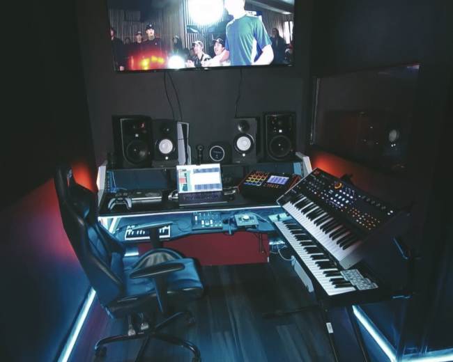 A music production studio features a desk with a keyboard, electronic instruments, and studio monitors, illuminated by LED lights, with a large screen displaying a performance.