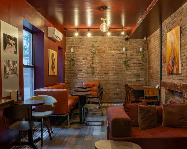 The image shows a cozy lounge area with brick walls, wooden furniture, and colorful artworks on display.
