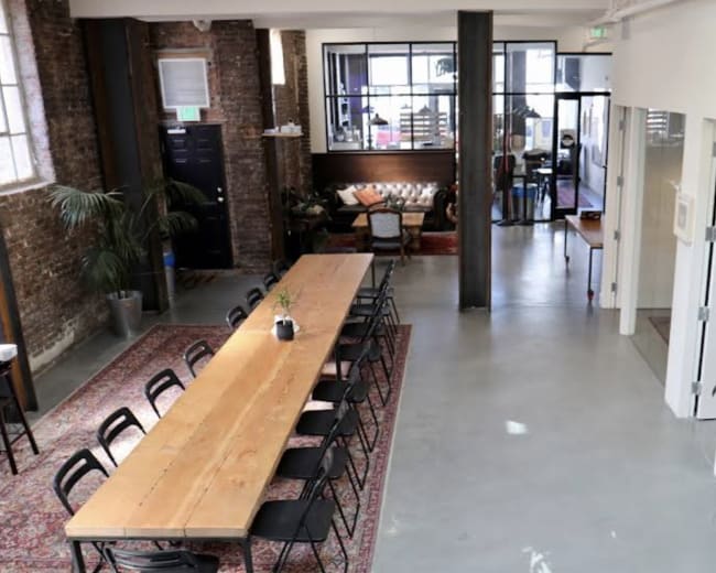 The image shows a spacious interior with a long wooden table surrounded by chairs, a seating area with couches, and large windows allowing natural light to enter.