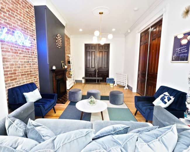 A cozy living room features a mix of modern furniture and exposed brick walls, with blue sofas arranged around a circular coffee table.