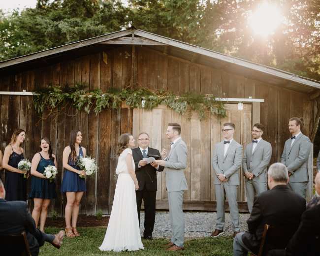 A couple exchanges vows during an outdoor wedding ceremony in front of a rustic wooden backdrop, surrounded by bridal party members and guests.