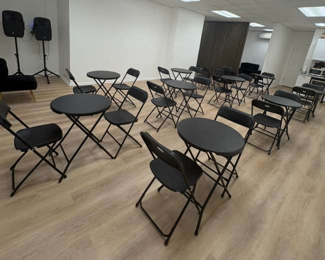 The image shows a room arranged with multiple circular tables and black folding chairs set up in a grid pattern.